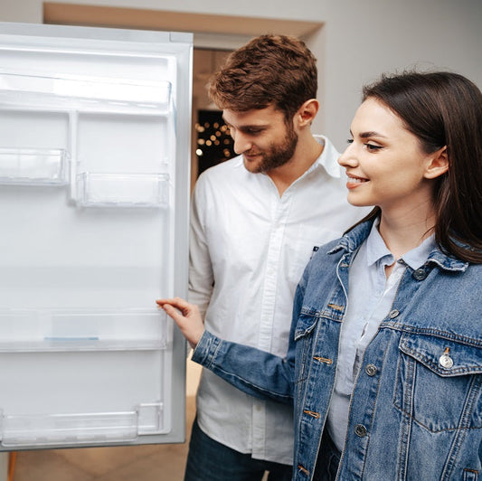 A young couple smiling while inspecting the open door shelves of a new refrigerator inside an appliance store.
